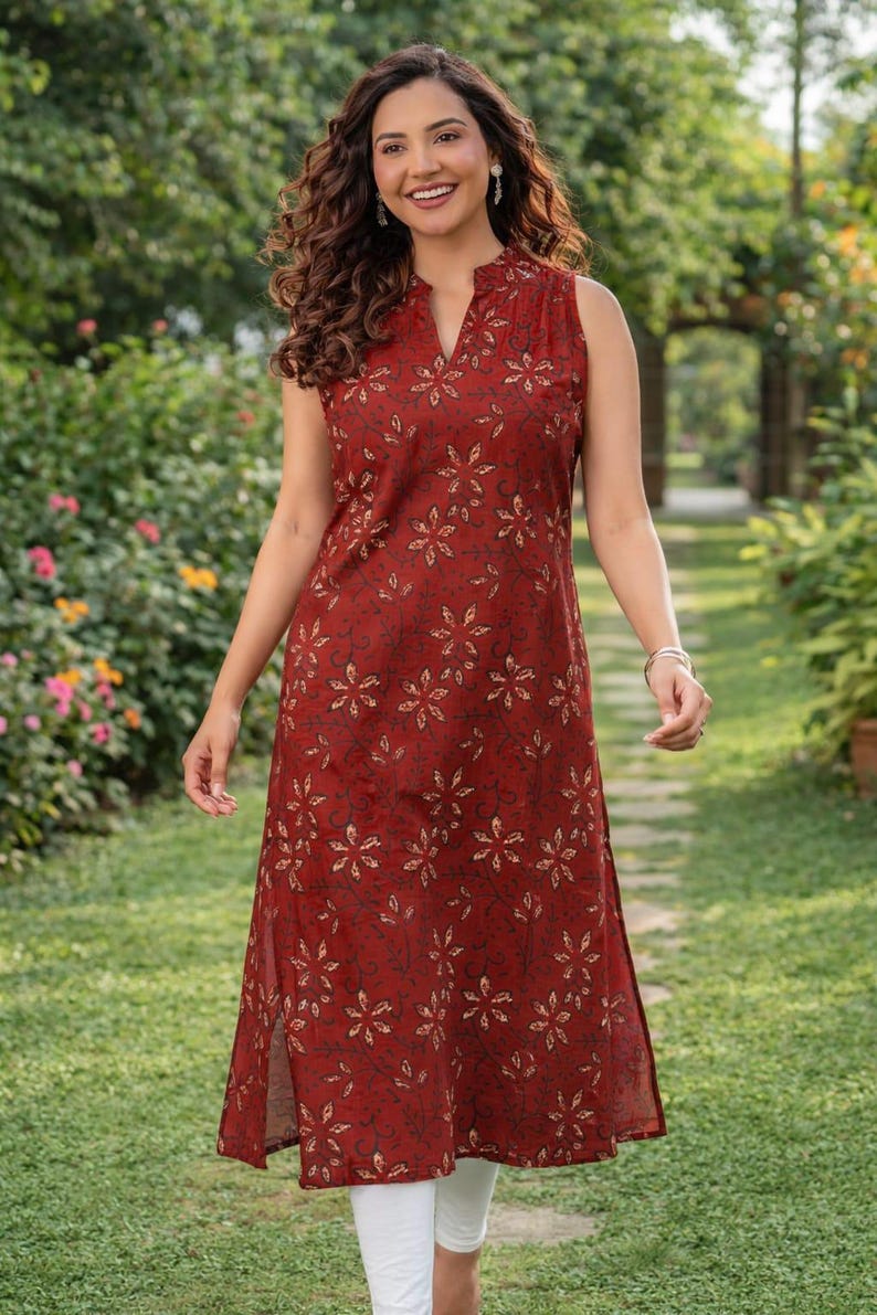 May include: A sleeveless, knee-length dress in a deep red hue, adorned with a repeating floral pattern in a lighter shade. The dress features a mandarin collar and side slits. The model is wearing white leggings.