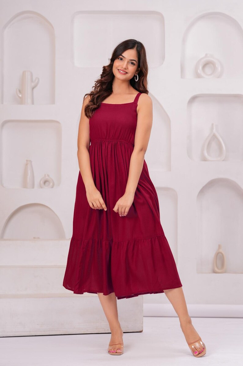 May include: A burgundy midi dress with a square neckline and spaghetti straps. The dress features a gathered waist and a tiered skirt. The model is wearing clear sandals.