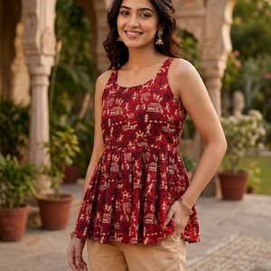 May include: A red sleeveless top with a flared hem, featuring a repeating pattern of gold and black figures and buildings. The top is worn with tan trousers. The style is casual and ethnic.