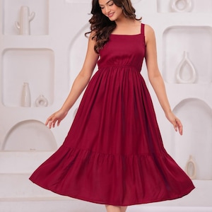 May include: A burgundy-colored dress with a square neckline and wide straps. The dress features a gathered bodice and a tiered, flowing skirt. The model is smiling and wearing the dress in a studio setting.
