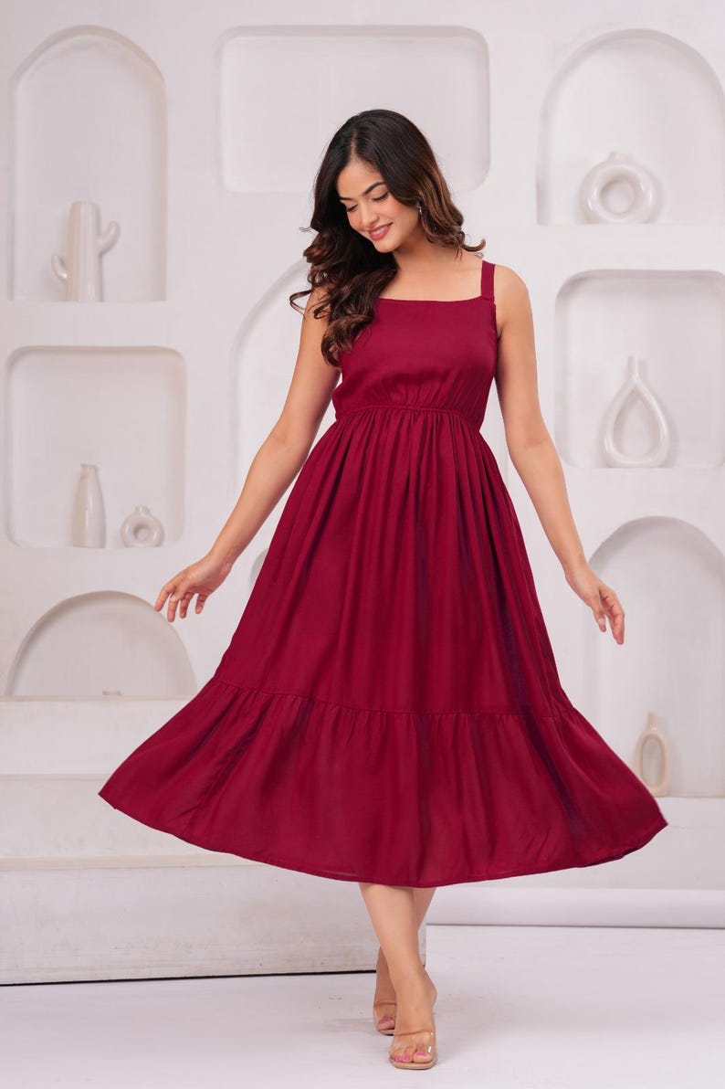 May include: A burgundy-colored dress with a square neckline and wide straps. The dress features a gathered bodice and a tiered, flowing skirt. The model is smiling and wearing the dress in a studio setting.