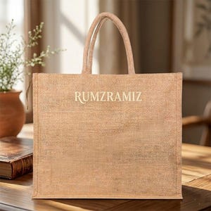 May include: A tan jute tote bag with the word "RUMZRAMIZ" printed in a light color. The bag has two handles and is sitting on a wooden surface. A small plant in a brown pot is visible in the background.