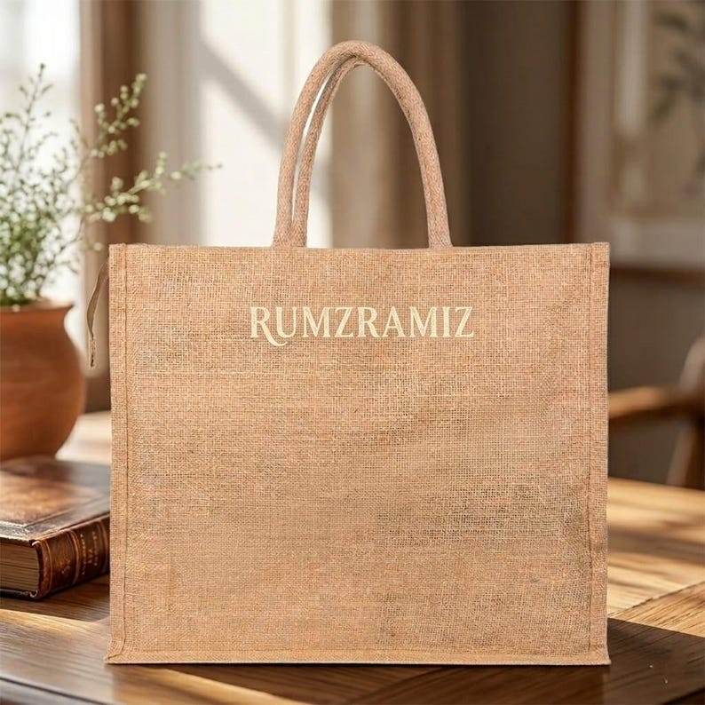 May include: A tan jute tote bag with the word "RUMZRAMIZ" printed in a light color. The bag has two handles and is sitting on a wooden surface. A small plant in a brown pot is visible in the background.
