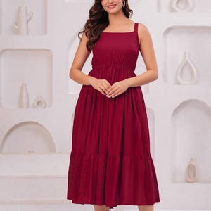 May include: A burgundy midi dress with a square neckline and wide straps. The dress features a fitted bodice and a tiered skirt. The model is wearing clear heels. The background is a white wall with decorative alcoves.