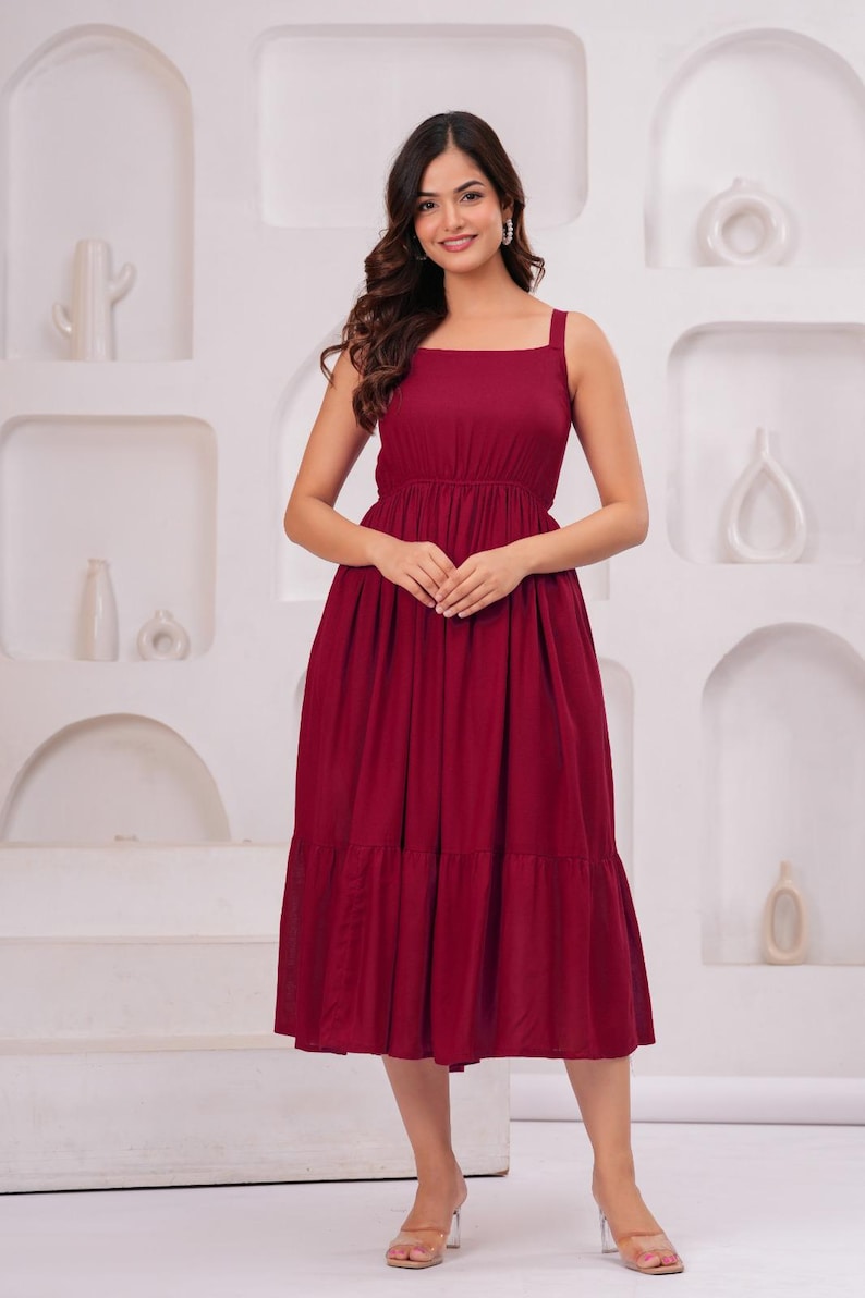 May include: A burgundy midi dress with a square neckline and wide straps. The dress features a fitted bodice and a tiered skirt. The model is wearing clear heels. The background is a white wall with decorative alcoves.
