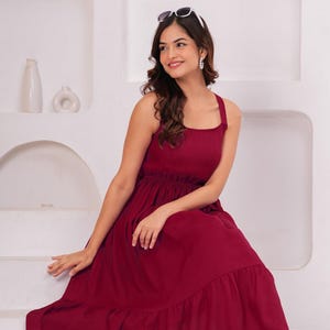 May include: A sleeveless, burgundy-colored maxi dress with a square neckline and a tiered skirt. The dress features a fitted bodice and a flowing skirt. Sunglasses are perched atop the head. The dress is suitable for casual or semi-formal occasions.
