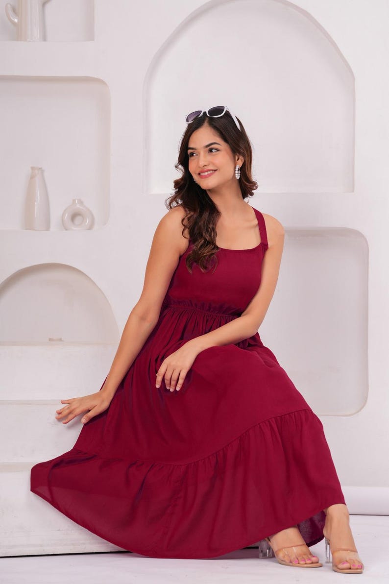 May include: A sleeveless, burgundy-colored maxi dress with a square neckline and a tiered skirt. The dress features a fitted bodice and a flowing skirt. Sunglasses are perched atop the head. The dress is suitable for casual or semi-formal occasions.