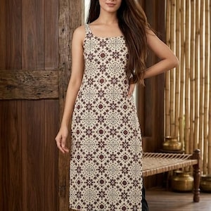 May include: A sleeveless, cream-colored tunic dress with a repeating maroon floral pattern. The dress is paired with black pants. The background features wooden paneling and a bench.