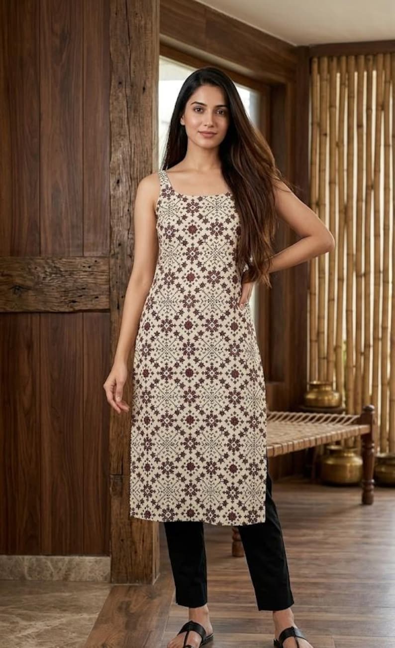 May include: A sleeveless, cream-colored tunic dress with a repeating maroon floral pattern. The dress is paired with black pants. The background features wooden paneling and a bench.