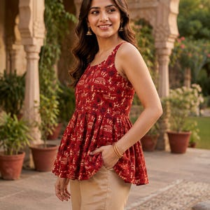 May include: A red sleeveless peplum top with a repeating pattern of small figures and buildings, paired with tan trousers. The woman wears gold earrings and bracelets. The background shows potted plants and architectural details.