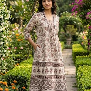 May include: A beige and brown floral print dress with three-quarter sleeves and pockets. The dress features a decorative border at the hem and a tassel detail at the neckline. The woman is wearing flat sandals and is standing in a garden.