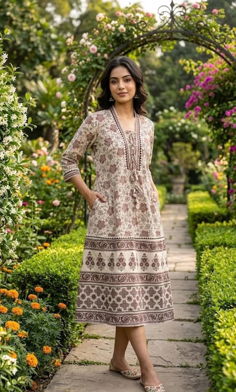 May include: A beige and brown floral print dress with three-quarter sleeves and pockets. The dress features a decorative border at the hem and a tassel detail at the neckline. The woman is wearing flat sandals and is standing in a garden.