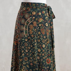 Puede incluir: Una camiseta sin mangas blanca combinada con una falda cruzada de color verde oscuro con un estampado floral detallado y un borde inferior que muestra un motivo repetitivo de elefantes rojos y dorados. La falda incluye un lazo lateral.