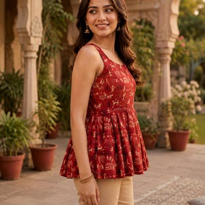 May include: A red sleeveless top with a flared peplum hem and a cream-coloured pattern. The top is paired with tan trousers. The top's pattern includes figures and shapes. The woman is wearing gold earrings and bracelets.