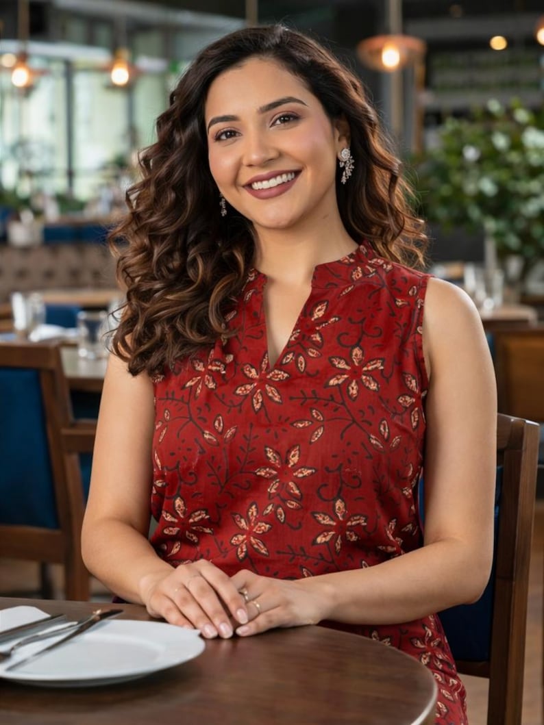 May include: A woman wearing a sleeveless, red top with a floral pattern. The top has a V-neck and is paired with a matching skirt. She is seated at a table in a restaurant, smiling at the camera. The background includes tables, chairs, and decorative lighting.