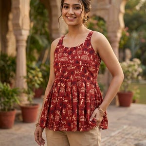 May include: A red sleeveless top with a distinctive pattern of small figures and shapes. The top features a pleated, flared design and is worn with tan trousers. The woman is wearing gold earrings and bracelets.