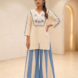 Könnte beinhalten: Eine cremeweiße Tunika mit V-Ausschnitt, blauer Blumenstickerei und blauen Quasten an den Ärmeln und am Oberteil, kombiniert mit einer weiten Hose mit vertikalen blauen und cremeweißen Streifen sowie cremeweißen, spitz zulaufenden Schuhen.