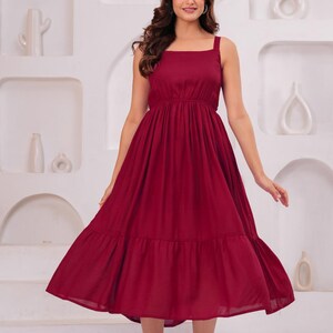 May include: A burgundy midi dress with a square neckline and wide straps. The dress features a fitted bodice and a flowing, tiered skirt. The model is wearing clear heels. The background is a white wall with decorative alcoves.