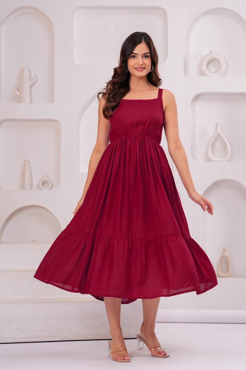 May include: A burgundy midi dress with a square neckline and wide straps. The dress features a fitted bodice and a flowing, tiered skirt. The model is wearing clear heels. The background is a white wall with decorative alcoves.