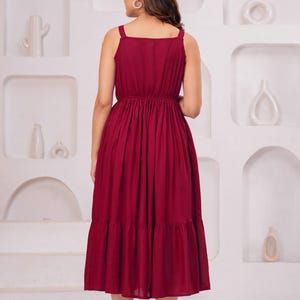 May include: A burgundy midi dress with a square neckline and spaghetti straps. The dress features a gathered waist and a tiered skirt. The model is wearing clear heeled sandals. The background is a white wall with decorative alcoves.