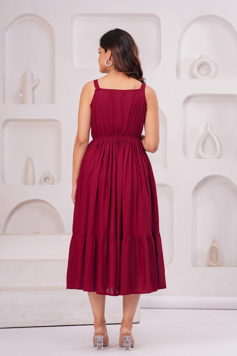 May include: A burgundy midi dress with a square neckline and spaghetti straps. The dress features a gathered waist and a tiered skirt. The model is wearing clear heeled sandals. The background is a white wall with decorative alcoves.