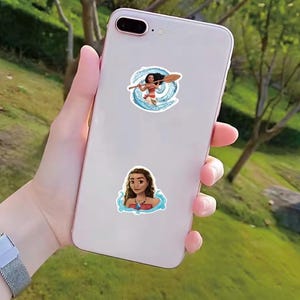 Puede incluir: Dos pegatinas con un personaje de dibujos animados de la pel&iacute;cula Moana se muestran en un tel&eacute;fono inteligente blanco. Una pegatina muestra al personaje con una paleta, rodeado de agua azul. La otra pegatina muestra al personaje con un collar.