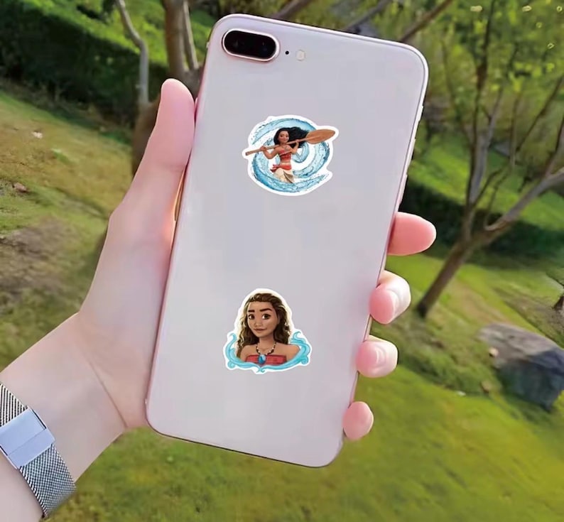 Puede incluir: Dos pegatinas con un personaje de dibujos animados de la pel&iacute;cula Moana se muestran en un tel&eacute;fono inteligente blanco. Una pegatina muestra al personaje con una paleta, rodeado de agua azul. La otra pegatina muestra al personaje con un collar.
