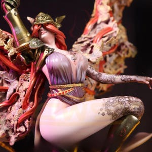 Könnte beinhalten: Detaillierte Figur einer Frau in einem fließenden lila und goldenen Kleid, verziert mit aufwendigen Blumen- und Blattmustern. Die Figur hat lange rote Haare, einen goldenen Helm und detaillierte Tätowierungen an Arm und Bein. Die Figur ist dynamisch posiert.