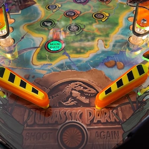 Könnte beinhalten: Nahaufnahme eines Jurassic Park Flipperautomaten. Das Spielfeld zeigt ein Kartendesign mit einer Dinosauriergrafik und dem Text "Jurassic Park". Die Flipper sind orange und gelb mit schwarzen Streifen.