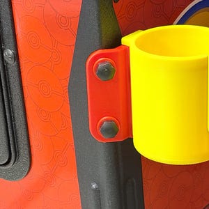 Può includere: Un portabicchieri cilindrico giallo brillante con un'apertura laterale verticale, fissato al telaio di un cabinato da gioco rosso e nero tramite una staffa di montaggio rossa fissata con due bulloni esagonali scuri. Il mobile presenta un motivo rosso di icone circolari.