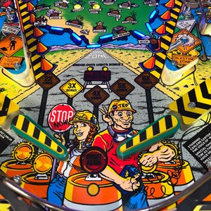 Könnte beinhalten: Ein farbenfrohes Flipper-Spielfeld mit Straßenbau-Thema. Enthält Zeichentrickfiguren, Straßenschilder und den Text "STOP NOW". Enthält Bonusmultiplikatoren und Städtenamen.