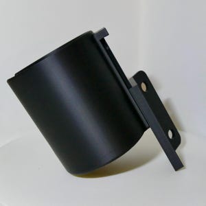 Op de afbeelding: Een zwarte, cilindrische bekerhouder met een montagebeugel. De bekerhouder is schuin en heeft een rechthoekige lip aan de bovenkant. De beugel heeft twee gaten voor montage. Het product is gemaakt van matzwart materiaal.