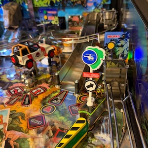 Könnte beinhalten: Ein Flipperautomat mit Jurassic Park-Thema. Das Spielfeld zeigt einen Miniatur-Jeep in Weiß und Orange, einen grünen und gelben Stoßfänger sowie Schilder mit der Aufschrift "HELIPAD" und "RESCUE". Der Hintergrund zeigt eine Dschungelszene.