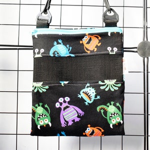 May include: A black fabric pouch with a zipper closure and a mesh pocket. The pouch has a colorful pattern of cartoon monsters on a black background. The pouch is hanging from a black adjustable strap.
