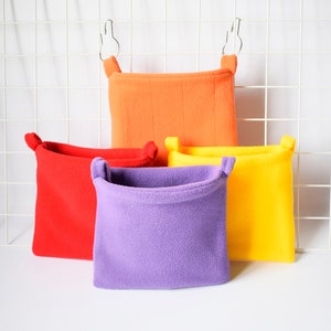 Pode incluir: Quatro camas de estimação de lã penduradas em diferentes cores: vermelho, laranja, amarelo e roxo. As camas são feitas de tecido macio e felpudo e têm um laço na parte superior para pendurar.