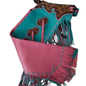 Peut inclure: Une paire de jambières chaps présentant un coloris turquoise et rose vif. La partie supérieure comprend du cuir marron avec des motifs floraux gravés, tandis que la partie inférieure est rose uni avec de longues franges roses et turquoise.