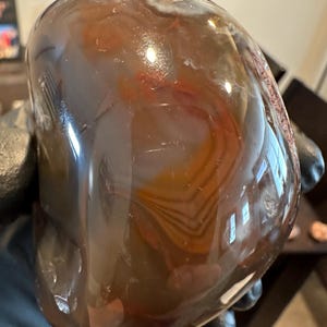 May include: A polished, rounded stone with a mix of brown, orange, and white swirling patterns. The stone has a smooth, glossy surface, reflecting light and showcasing the natural variations in color and texture. The stone is a decorative item.