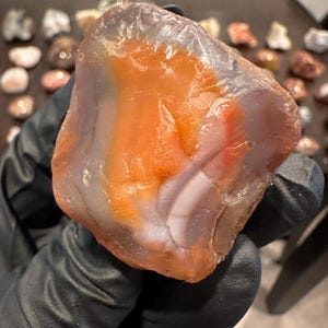 May include: A polished, translucent agate stone featuring vibrant bands of orange, white, and soft gray. The mineral specimen has a smooth, rounded surface with visible internal layering and a glossy, semi-transparent texture throughout the rock.