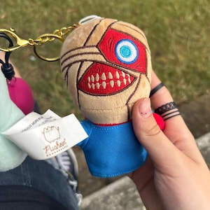 以下が含まれることがあります： ベージュの頭、赤いアクセント、青いシャツを着たキャラクターのぬいぐるみキーホルダー。キャラクターは、白い歯と青と白の目を持つ大きな赤い口をしています。ゴールドカラーのキーホルダーと「Pusheen」という言葉のタグが付属しています。