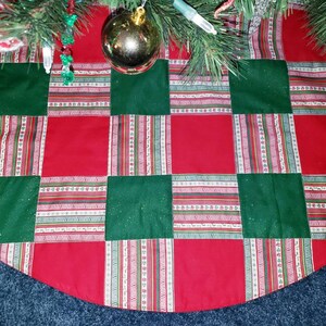 May include: A Christmas tree skirt with a patchwork design. The skirt features alternating red and green squares. The red squares are solid, while the green squares have a subtle sparkle. Decorative striped ribbons with white, red, and green accents are sewn between the squares.