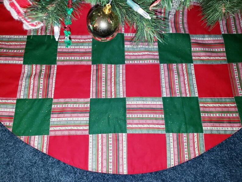 May include: A Christmas tree skirt with a patchwork design. The skirt features alternating red and green squares. The red squares are solid, while the green squares have a subtle sparkle. Decorative striped ribbons with white, red, and green accents are sewn between the squares.