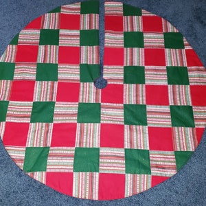 May include: A round Christmas tree skirt with a patchwork design. The skirt features a pattern of red and green squares, with striped accents in between. The skirt has a split opening and a hole in the center for the tree trunk.