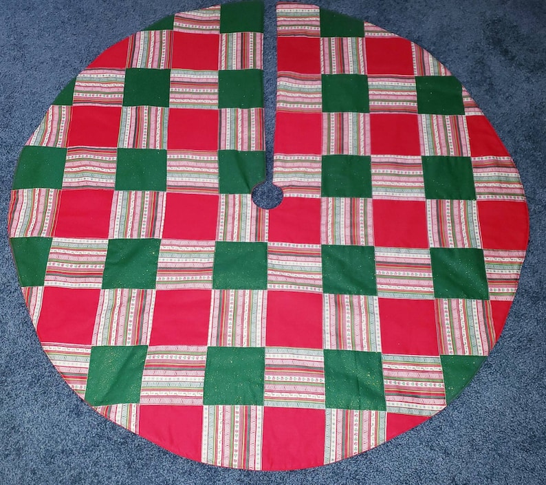 May include: A round Christmas tree skirt with a patchwork design. The skirt features a pattern of red and green squares, with striped accents in between. The skirt has a split opening and a hole in the center for the tree trunk.