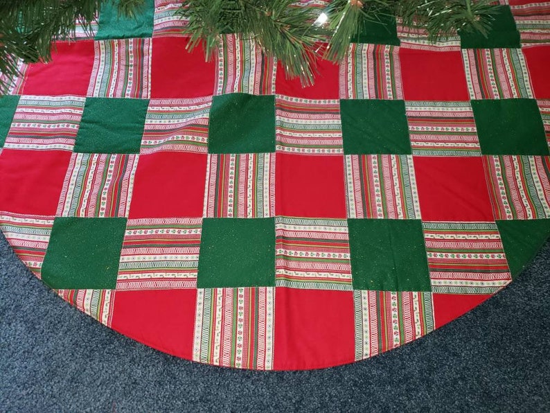 May include: A Christmas tree skirt with a patchwork design. The skirt features alternating red and green squares, with striped fabric accents in between. The skirt is made of fabric and has a semi-circular shape.