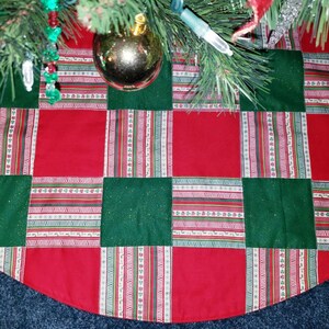 May include: A quilted Christmas tree skirt with a patchwork design. The skirt features alternating red and green squares, with a striped pattern in between. The skirt is under a Christmas tree with ornaments.
