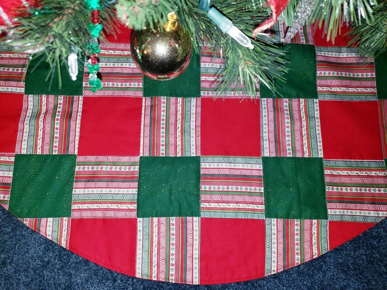 May include: A quilted Christmas tree skirt with a patchwork design. The skirt features alternating red and green squares, with a striped pattern in between. The skirt is under a Christmas tree with ornaments.