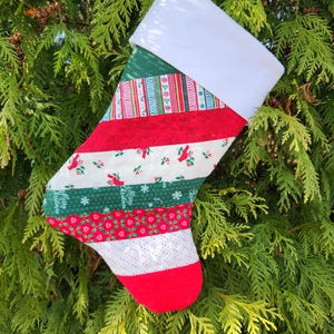 May include: A handmade Christmas stocking with a white cuff and a red hanging loop. The body features various patterned fabric strips in red, green, and white, with festive designs like snowflakes and birds. The toe is solid red.