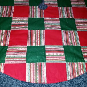 May include: A Christmas tree skirt with a patchwork design. The skirt features alternating red and green squares, with patterned stripes in between. The skirt has a scalloped edge and a hole in the center for the tree trunk.