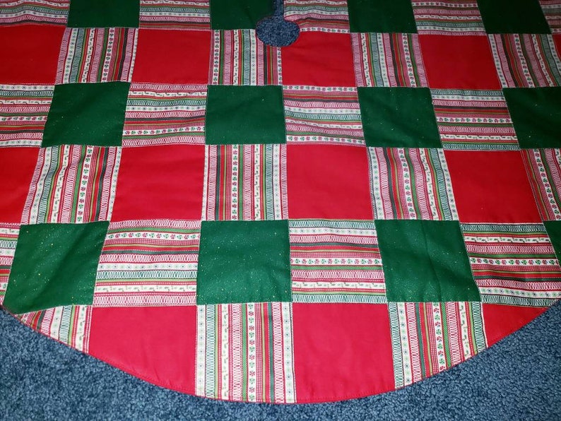 May include: A Christmas tree skirt with a patchwork design. The skirt features alternating red and green squares, with patterned stripes in between. The skirt has a scalloped edge and a hole in the center for the tree trunk.