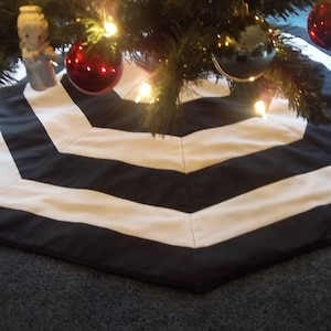 May include: A black and white chevron patterned Christmas tree skirt. The skirt is hexagonal in shape and made of fabric. Red and black ornaments and a small angel figurine decorate the tree.